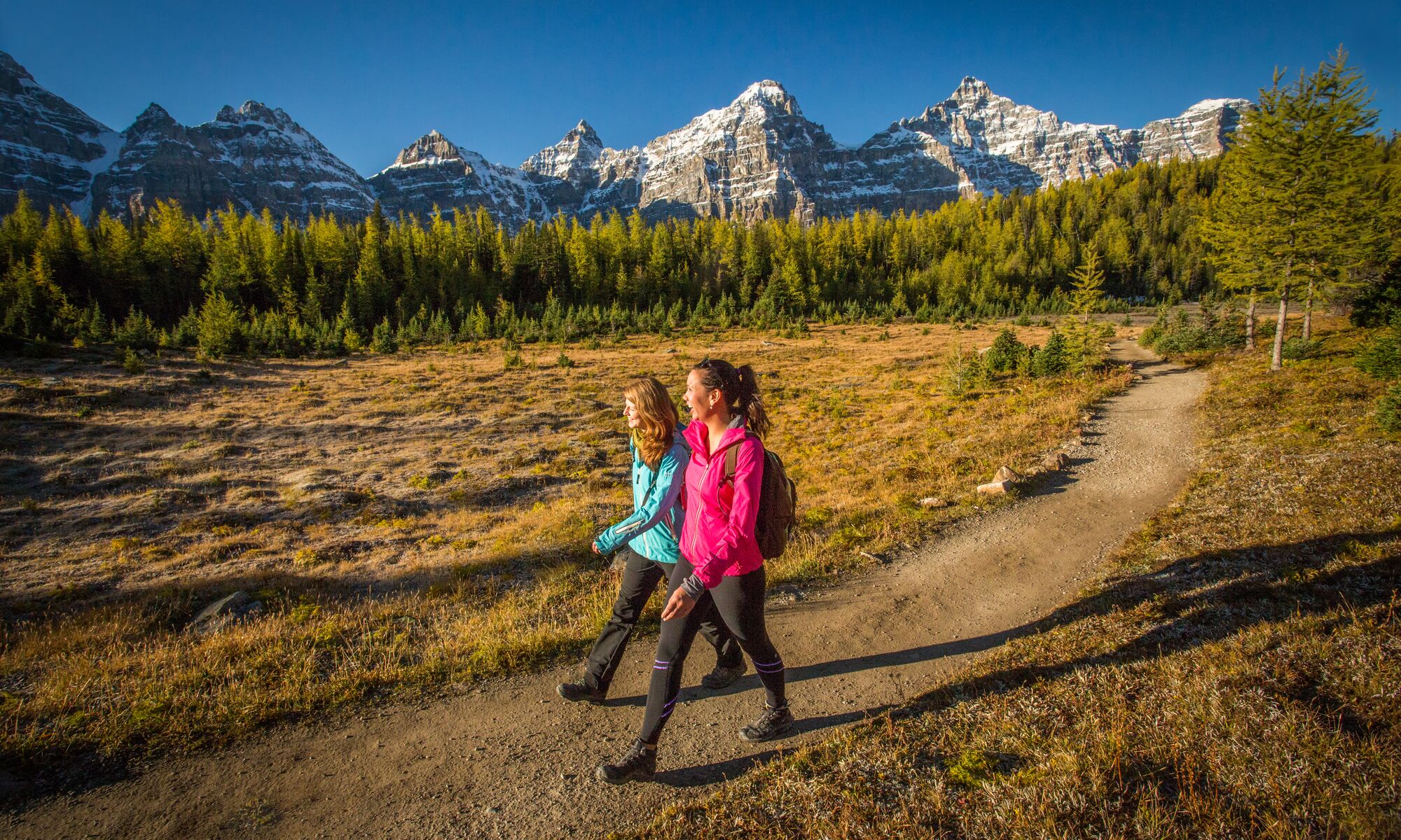 Two women walk in Larch Valley with mountain peaks behind them in Banff National Park. 